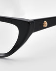 Close-up of the corner of a black cat-eye optical frame featuring a gold rose emblem on the arm, from For Art's Sake® Rosalia II, against a plain gray background.