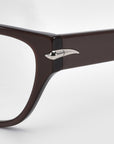 Close-up of For Art's Sake® Eros cat-eye acetate frames in brown, featuring a silver accent on the temple hinge. These chic sunglasses offer a sleek and modern look against a light gray background, making them perfect for everyday wear.