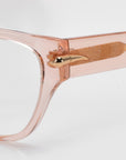 Close-up of Eros by For Art's Sake®: translucent cat-eye acetate frames in light pink, featuring gold metallic horn detail on the hinges. Ideal for everyday wear, these sunglasses add elegance and perfectly complement a rose garden backdrop.