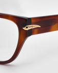 Close-up of the Eros eyeglass frames by For Art's Sake®. These brown, rectangular frames feature a unique gold metallic horn-shaped detail on the hinge against a plain white background, highlighting their elegant design.