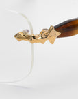 Close-up of For Art's Sake® Claremont eyeglasses; rimless rectangular lenses with dark brown arms and gold geometric designs on the temples from the Rose Garden collection.