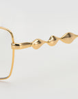 Close-up of For Art's Sake® Fluid gold eyeglasses with a soft-square silhouette and unique, wavy sculpted arms. The thin metallic frame highlights undulating temples, showcasing the brand’s signature fluid optical design against a white background.