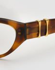 Close-up of the side of the Halo(X) sculptural brown tortoiseshell acetate frame by For Art's Sake®, featuring two gold metal accents on the temple against a light gray background.