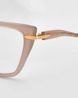 Close-up of the hinge on Motion I cat-eye sunglasses by For Art's Sake®, featuring a light brown translucent frame, gold accents, and acetate temples against a plain light background.