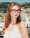 A woman with long, wavy blonde hair wears For Art's Sake® Motion II glasses with acetate temples and a white tank top, standing before yachts and buildings on a sunny day, hills in the background.