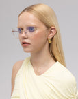 A woman with long blonde hair wears For Art's Sake® Garden squared metal opticals, gold hoop earrings, and a light yellow asymmetrical top against a plain light gray background.