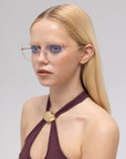 A young woman with long blonde hair wears For Art's Sake® Garden modern squared metal glasses and a sleeveless purple top with a gold clasp, gazing slightly to the side against a light gray background.