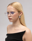 A young woman with straight blonde hair wears For Art's Sake® Garden modern squared metal glasses, silver geometric earrings, and a black off-shoulder top with a decorative brooch, gazing to the side against a plain background.