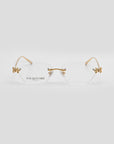 The For Art's Sake® Ida eyeglasses are rimless and lightweight with gold temples and butterfly-shaped corner accents. The left lens features the brand name, while a subtle floral design decorates the light gray background.