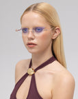 A woman with straight blonde hair models For Art's Sake® Ida rimless cat-eye glasses with purple-tinted lenses, styled with a dark purple halter top and gold clasp, posing before a light gray background.
