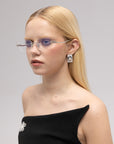 A woman with long blonde hair models For Art's Sake® Ida frameless glasses, bold eye makeup, geometric earrings, and a one-shoulder black dress with a jeweled flower, posing against a plain light background.