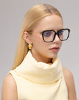 A woman with long blonde hair wears For Art's Sake® Claudia black-rimmed glasses with sculpted temples, gold earrings, and a sleeveless pale yellow high-collared top, looking slightly to the side against a neutral background.