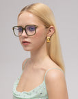A young woman with long blonde hair wears For Art’s Sake® Claudia sunglasses and geometric gold earrings, paired with a light green polka dot dress, standing against a plain light background.