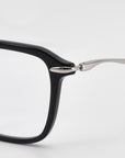 Close-up of the top left corner of For Art’s Sake® Claudia eyeglasses, showing a black rectangular acetate and metal frame with sculpted temples and silver hinge, set against a plain white background.