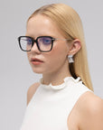 A young woman with straight blonde hair models the Claudia glasses by For Art's Sake®, featuring large black acetate and metal frames with sculpted temples, styled with geometric earrings and a sleeveless white ruffled top against a light background.