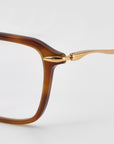 Close-up of the left side of For Art's Sake® Claudia eyeglasses, showing a brown rectangular acetate and metal frame with sculpted temples and a thin gold temple arm, set against a plain light gray background.