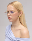 A young woman with straight blonde hair models the For Art's Sake® Anita geometric metal eyewear, paired with gold square earrings and a light blue off-shoulder top, against a neutral background.