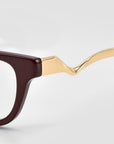 Close-up of For Art's Sake® Poppy eyeglasses featuring a dark brown front and a glossy, wavy gold sculptural metal temple, set against a light grey background.