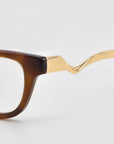 Close-up of For Art's Sake® Poppy eyeglasses with a brown rectangular frame and bold temples, featuring twisted gold sculptural metal temple arms against a light background.