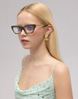 A woman with long blonde hair poses against a plain light background, wearing For Art's Sake® Lavender sculptural cat-eye acetate glasses, square gold earrings, and a light green sequined top.