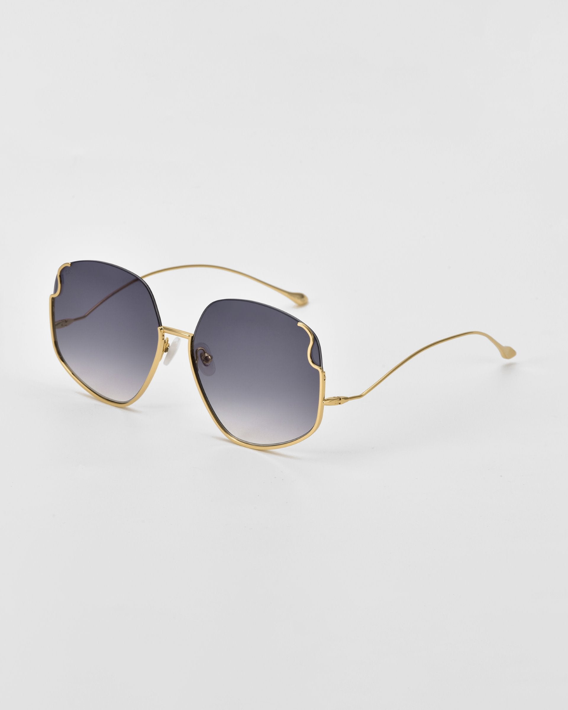 A pair of Drape sunglasses by For Art's Sake® with gold frames and dark, gradient lenses. The design features a unique, slightly wavy cut on the top edges of the oversized squared frame. Intricate metal detailing adds to their charm as they rest on a white surface, viewed at an angle.