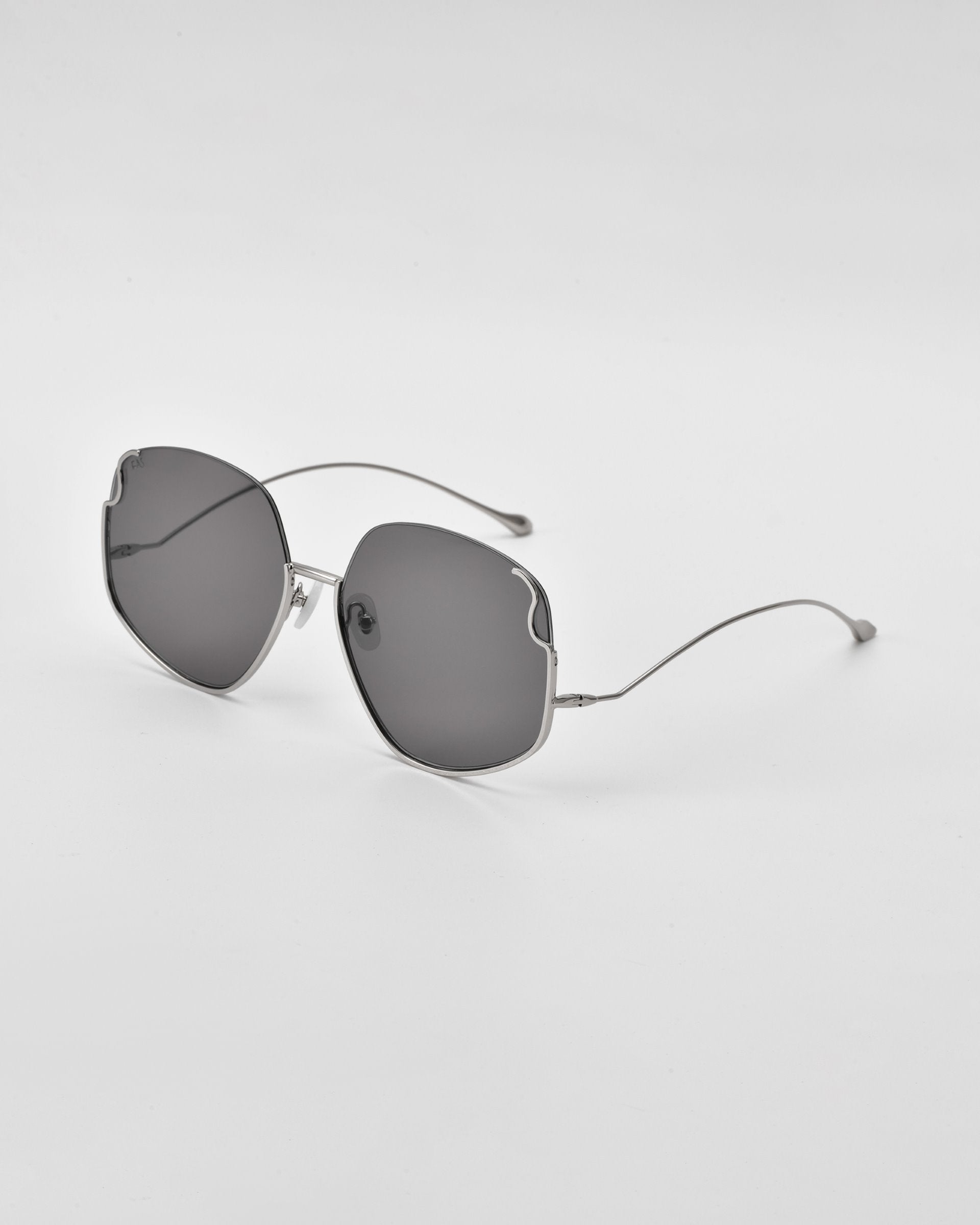 A pair of Drape sunglasses by For Art's Sake® with curved, wireframe arms and large, slightly rounded, dark lenses. The frames are fine and metallic with intricate metal detailing, creating a modern and minimalist look. The glasses are placed on a plain, light-colored surface.