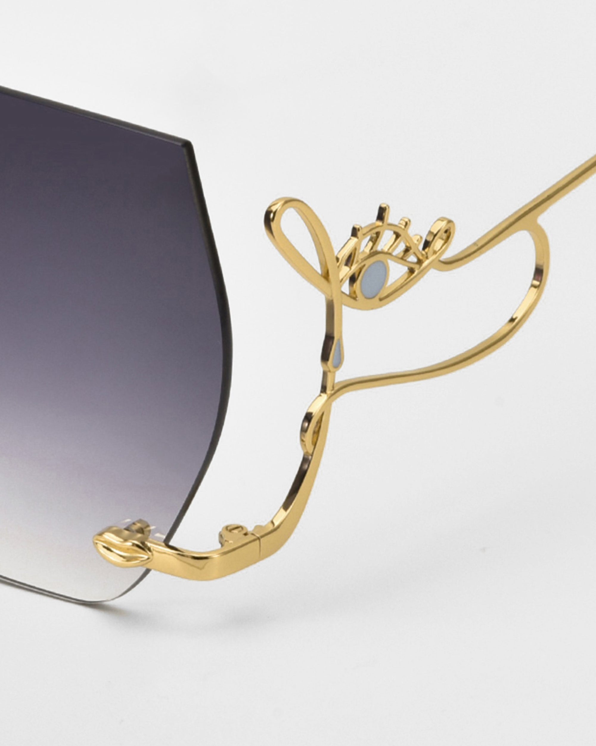 A close-up of For Art's Sake® Cry Me a River sunglasses shows a gold metal arm shaped like an eye with lashes and an artistic, dark angular lens that fades to clear at the bottom.