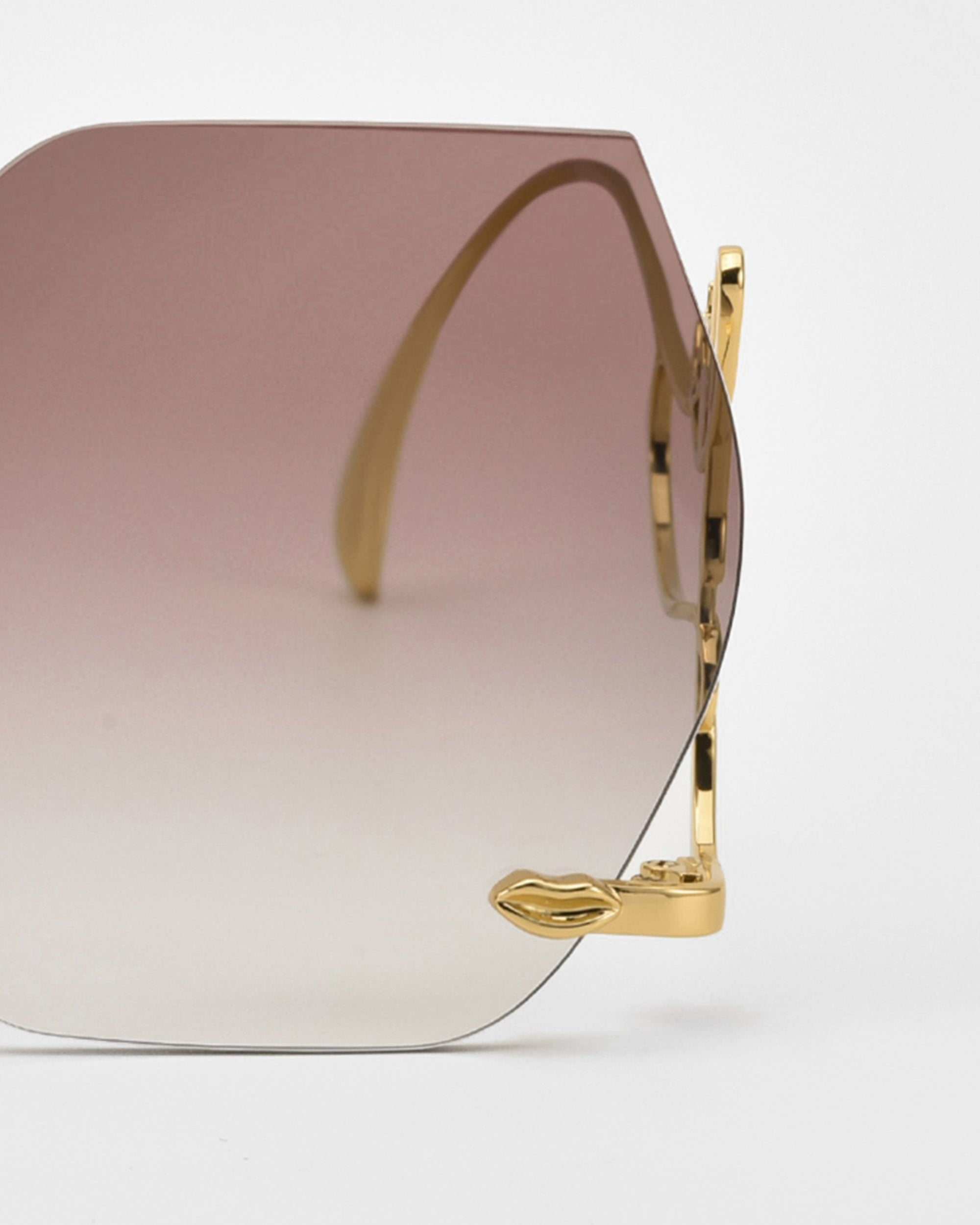 A close-up of For Art’s Sake® Cry Me a River sunglasses, featuring a gold frame with abstract details and a small gold lips-shaped accent on one corner, set against a plain white background.