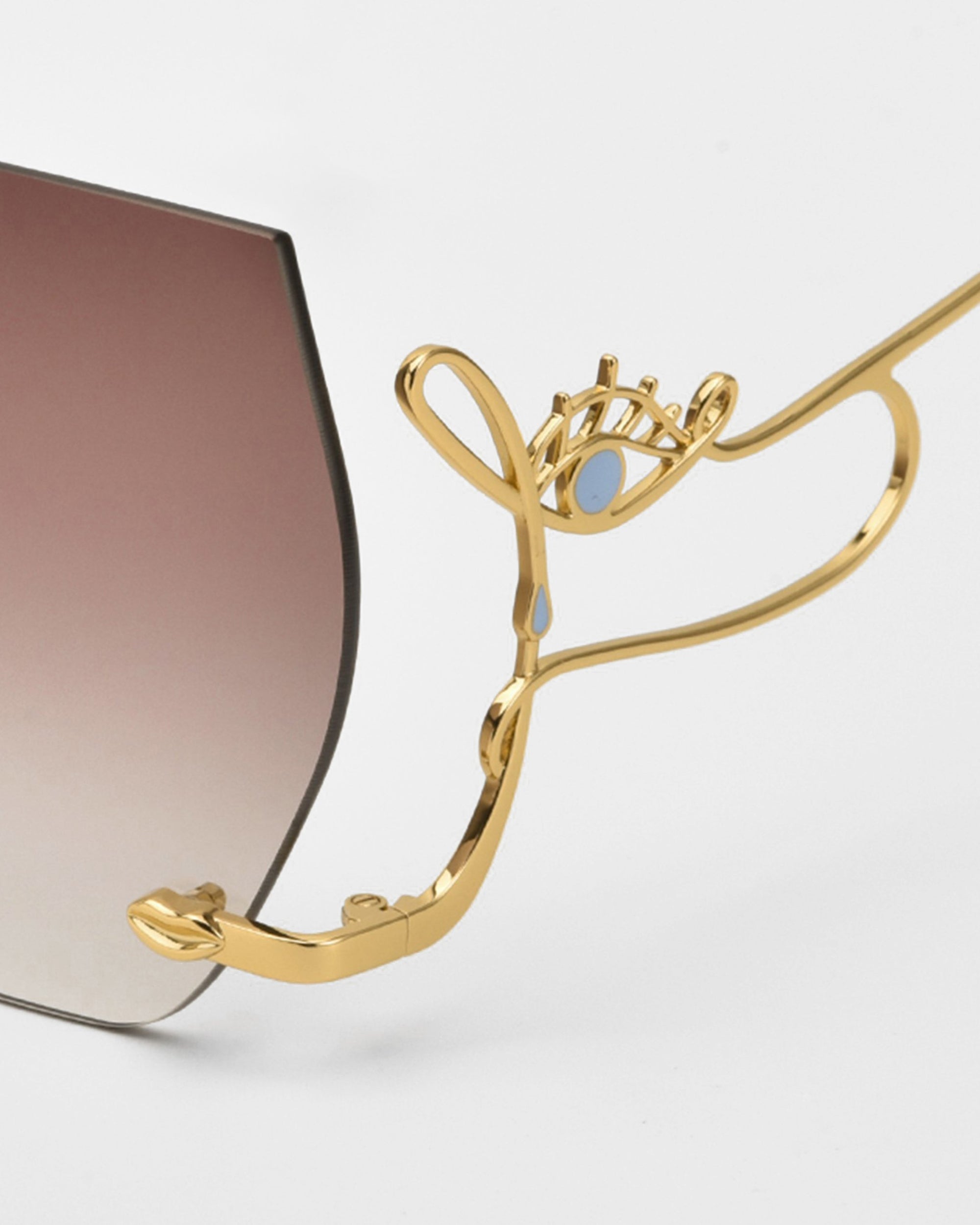 Close-up of For Art's Sake® Cry Me a River sunglasses with gold metal frames, abstract eye-shaped temples, blue accents, and gradient brown lenses—these limited edition statement shades stand out against a white background.