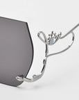 Close-up of For Art's Sake® Cry Me a River limited edition sunglasses, featuring dark lenses and an abstract silver metal eye-shaped arm with light blue enamel accents. A plain white background highlights their striking design.