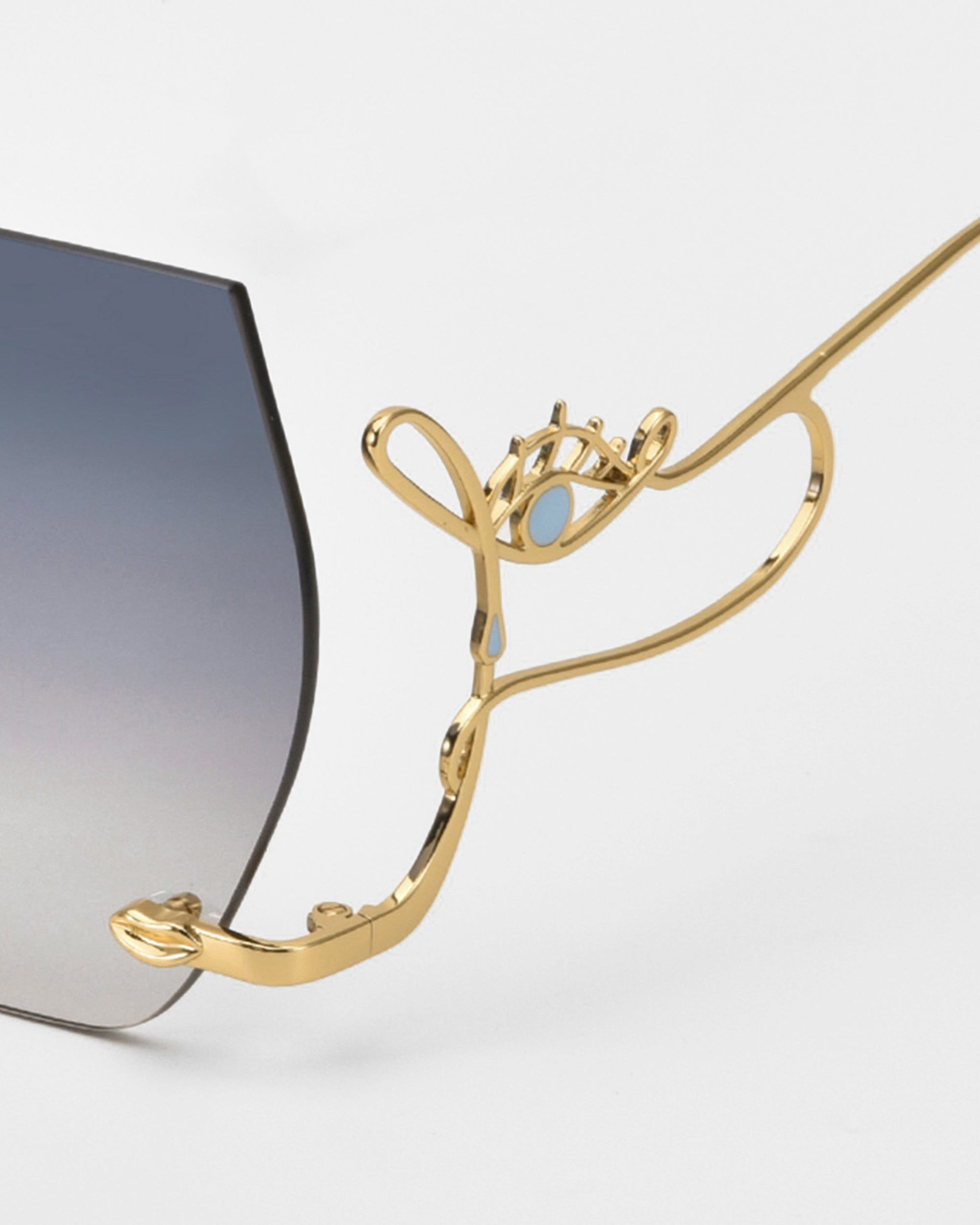 A close-up of For Art's Sake® Cry Me a River sunglasses shows a gold temple shaped like an abstract face and eye, attached to gradient dark-to-light gray lenses. The artistic sunglasses are displayed against a plain white background.
