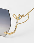 A close-up of For Art's Sake® Cry Me a River sunglasses shows a gold temple shaped like an abstract face and eye, attached to gradient dark-to-light gray lenses. The artistic sunglasses are displayed against a plain white background.