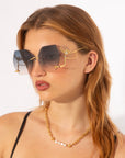 A person with long, light brown hair is wearing oversized, square-shaped statement sunglasses from For Art's Sake®'s limited edition Cry Me a River collection, featuring gradient dark lenses and gold accents. They also have a gold chain with pearls around their neck. The background is plain white.