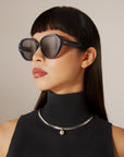A woman with straight black hair and blunt bangs wears For Art’s Sake® Mirage White plant-based acetate sunglasses, a sleeveless black turtleneck, and a silver pearl necklace as she gazes off to the side.