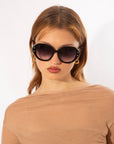 A woman with light brown hair wears large, round Mirage White sunglasses by For Art's Sake®, gold hoop earrings, and a sheer beige top. She has a neutral expression and stands against a plain white background.