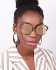 A person with dark skin and black hair styled in a top bun is wearing the Alectrona large circular glasses with tinted lenses from For Art's Sake®, 18kt gold-plated hoop earrings, and a white striped blazer. They are gazing directly at the camera against a plain white background.