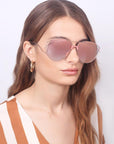 A woman with long, wavy brown hair is wearing the "Dark Eyes" sunglasses by For Art's Sake®. These large, reflective pink sunglasses feature stainless steel frames and Nylon lenses that provide 100% UV protection. She has a neutral expression and is dressed in a brown and white striped top, set against a plain white background.