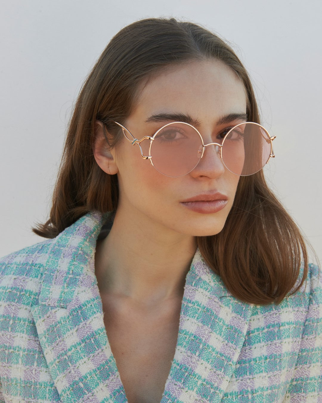 A woman with straight brown hair wears For Art's Sake® Diptych - Pink sunglasses (ONLY 2 LEFT), which feature UV protection, paired with a pastel plaid blazer as she looks past the camera against a plain light background.