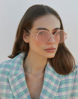 A woman with straight brown hair wears For Art's Sake® Diptych - Pink sunglasses (ONLY 2 LEFT), which feature UV protection, paired with a pastel plaid blazer as she looks past the camera against a plain light background.