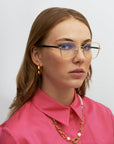 A person with long, light brown hair is wearing the Grace glasses from For Art's Sake®, featuring gold rims and handmade plant-based acetate frames. They are also sporting large gold hoop earrings and a pink collared shirt, showcasing a serious expression. Their look is completed with an 18-karat gold chain necklace adorned with white pearls against a plain white background.