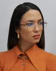 A person with straight black hair wears gold-rimmed Grace glasses from For Art's Sake®, adorned with a handmade frame of 18-karat gold, along with gold hoop earrings and an orange shirt featuring ribbed details, accessorized with a chain necklace. They have a thoughtful expression and look slightly to the side, set against a neutral gray background.