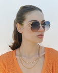 A woman with long brown hair wears For Art's Sake® Hera sunglasses (LAST ONE), featuring oversized UV-protective shades, a gold layered necklace, and an orange knit top, looking off to the side against a light background.