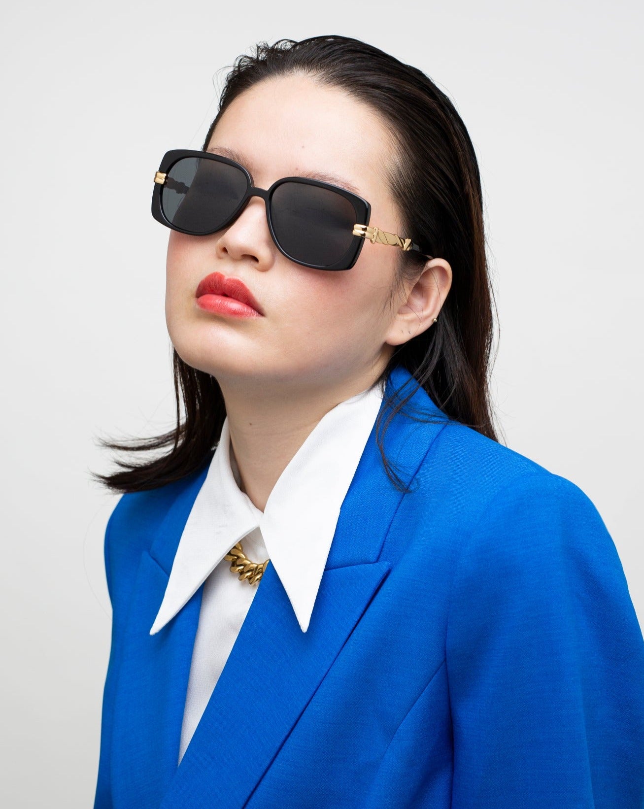 A person with long dark hair dons oversized Icon-ARC D-frame shades by For Art's Sake® and a blue blazer over a white shirt. The neutral expression is complemented by the sleek UVA & UVB-protected design, set against a gray background.