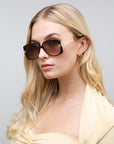 A person with long blonde hair wears Icon-ARC acetate sunglasses by For Art's Sake® and gold hoop earrings. The UVA & UVB-protected D-frame shades complement their yellow top as they glance to the side against a plain gray backdrop.