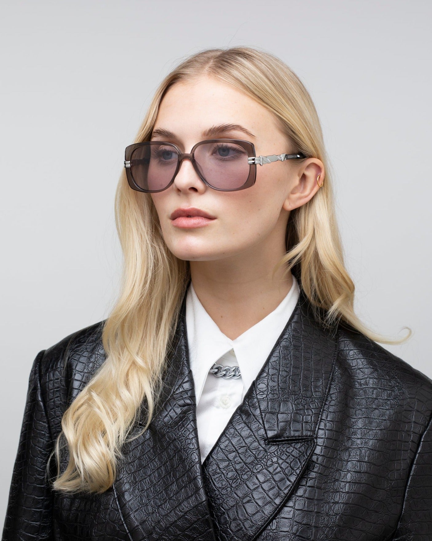 A person with long blonde hair dons For Art's Sake® Icon-ARC large handmade acetate sunglasses, a white collared shirt, and a black textured leather jacket. The neutral background accentuates their stylish look, while the D-frame shades offer full UVA & UVB protection.