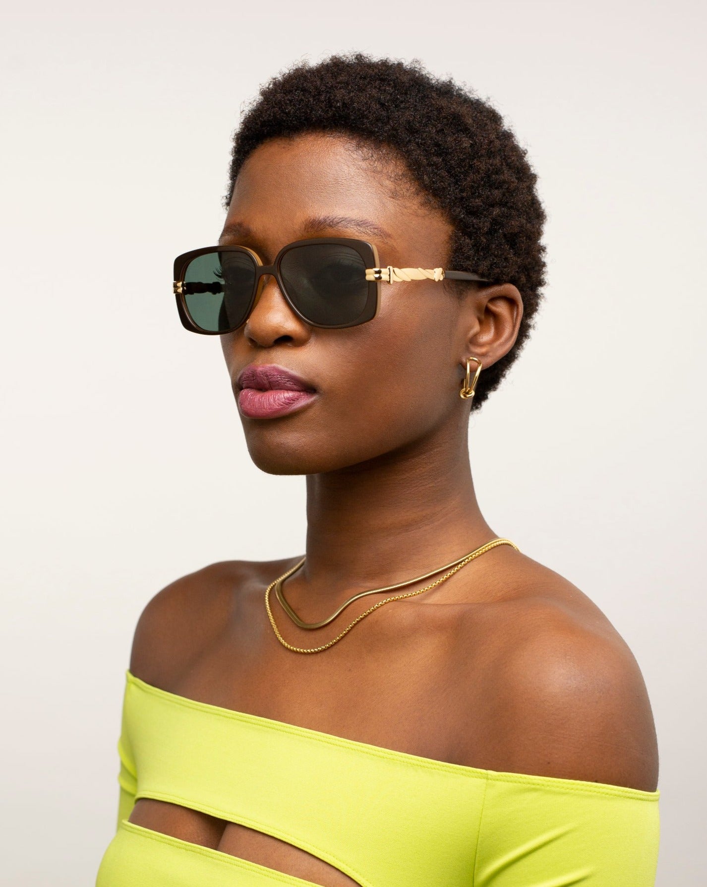 A person with a short hairstyle models handmade For Art's Sake® Icon-ARC acetate sunglasses against a neutral backdrop. The D-frame shades provide UVA & UVB protection and pair well with gold jewelry and a bright yellow off-the-shoulder top featuring cut-out details, enhancing the elegant style.