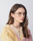 A woman with long brown hair and glasses from For Art's Sake® chic Lady-S collection wears a yellow knit cardigan with a pink stripe, a cream blouse with a bow, and gold hoop earrings styled like intricate twisted metalwork as she gazes to the side against a plain background.