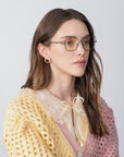 A woman with long brown hair, wearing For Art's Sake® Lady-S cat-eye glasses and hoop earrings, gazes sideways. She sports a ruffled light blouse under a pastel yellow and pink knitted cardigan against a plain, light-colored background.