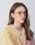 A woman with long brown hair and cat-eye silhouette glasses from For Art's Sake® looks to the side. She wears a Lady-S crochet yellow and pink sweater with a frilly collar and bow tie, her circular pink earrings charmingly contrasting the plain, light-toned background.