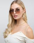 A woman with long blonde hair wears the Lemonade (Last Piece) statement sunglasses by For Art’s Sake®, paired with gold earrings and a gold necklace. She is dressed in an off-shoulder white ribbed top featuring thin straps and subtle faux-pearl detailing, set against a plain light background.
